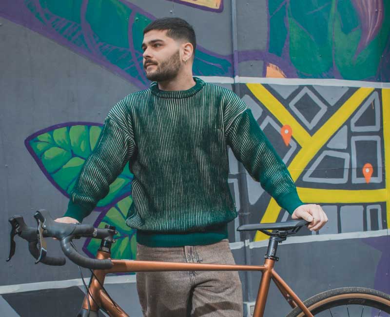 zamu-sweater-verde-ropa-hombre-bicicleta-estilo-urbano Hombre vistiendo sweater verde ZAMU junto a bicicleta urbana frente a mural colorido, vestuario funcional con estilo contemporáneo