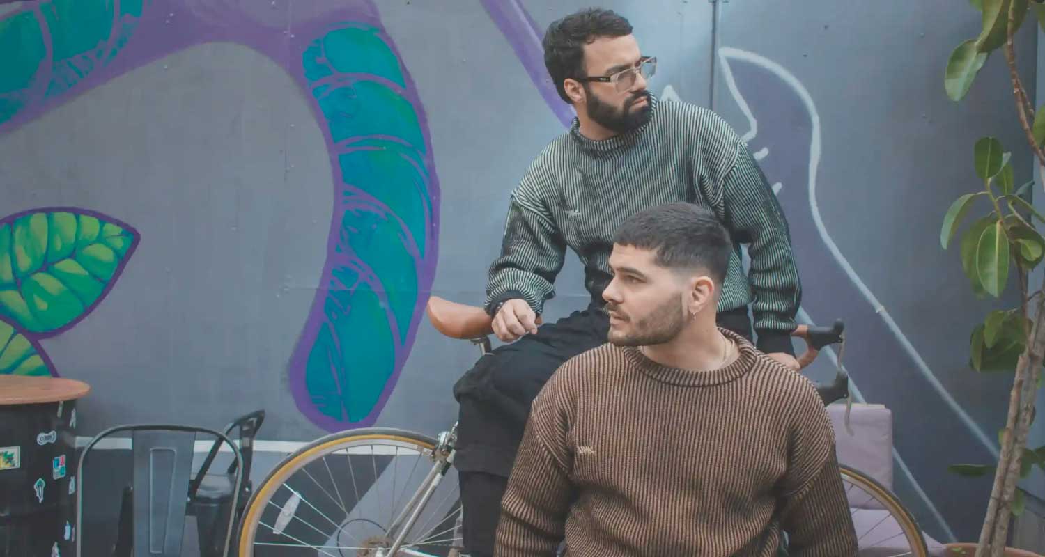 zamu-sweaters-diseno-chileno-hombres-estilo-urbano-bicicleta Dos hombres vistiendo sweaters ZAMU de diseño chileno en tonos tierra, frente a mural urbano con bicicleta vintage, vestuario masculino con identidad local